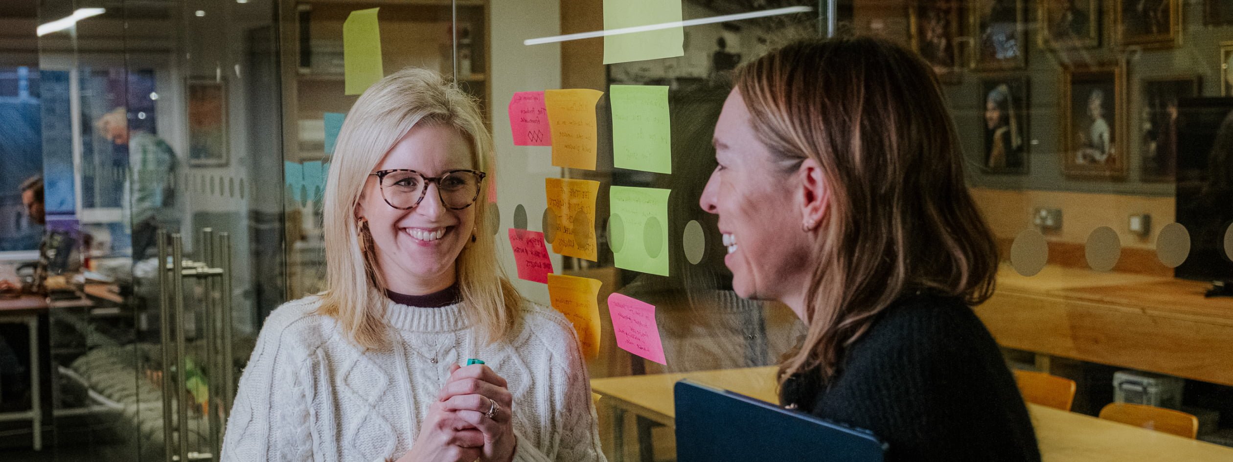 Two women smile and chat in an office, standing either side of a glass wall covered in colourful sticky notes.