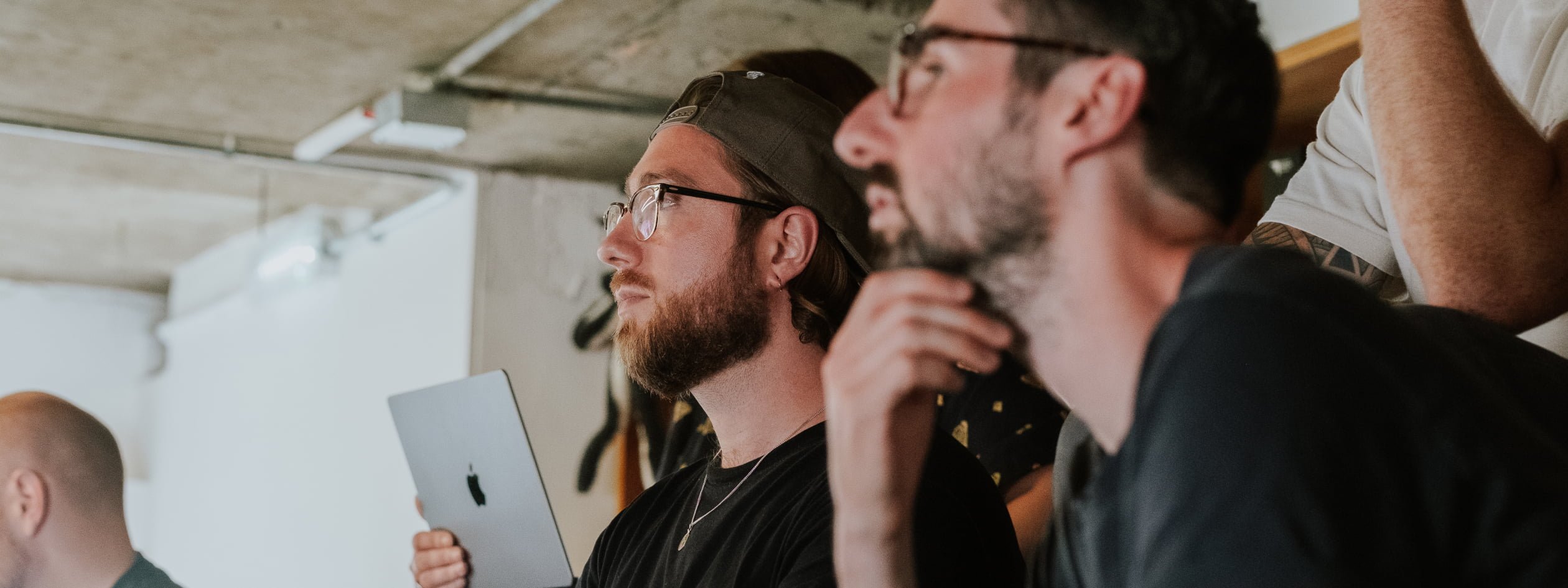 Two men look towards a presentation in a workshop setting, one holding a laptop, in a room with exposed concrete ceilings.