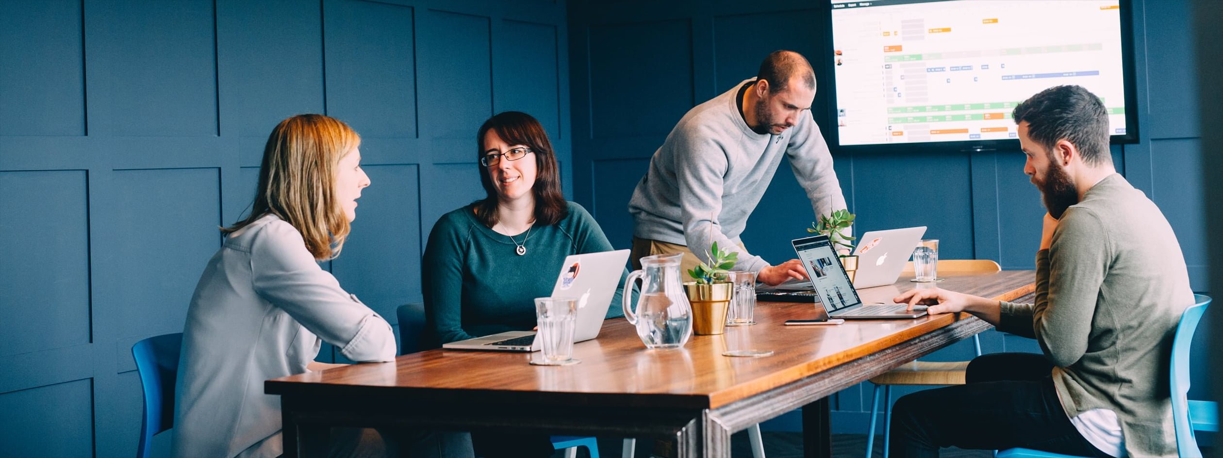 Four colleagues are gathered around a long table in a meeting space. Two are in conversation, while the other two are looking at their laptops. In the background a large screen mounted on a wall displays a detailed project schedule breakdown.