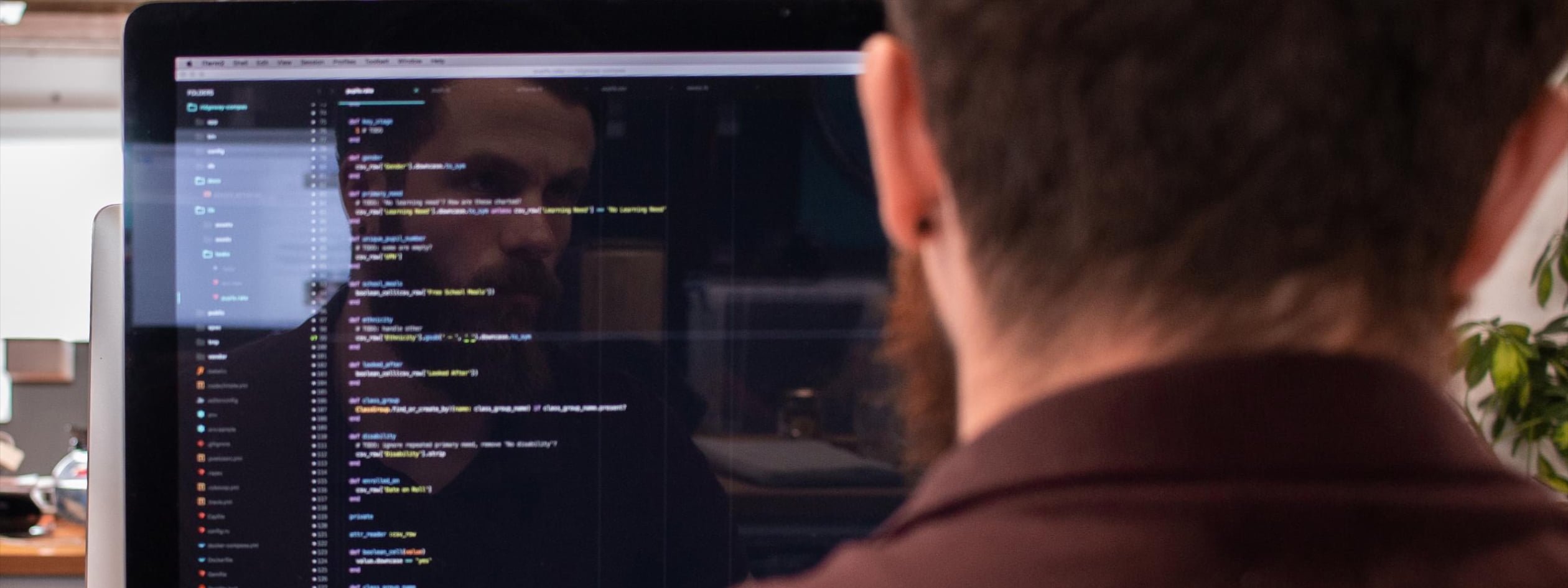 A close-up over the shoulder shot of a man working at a computer. On his screen is a code editor application.
