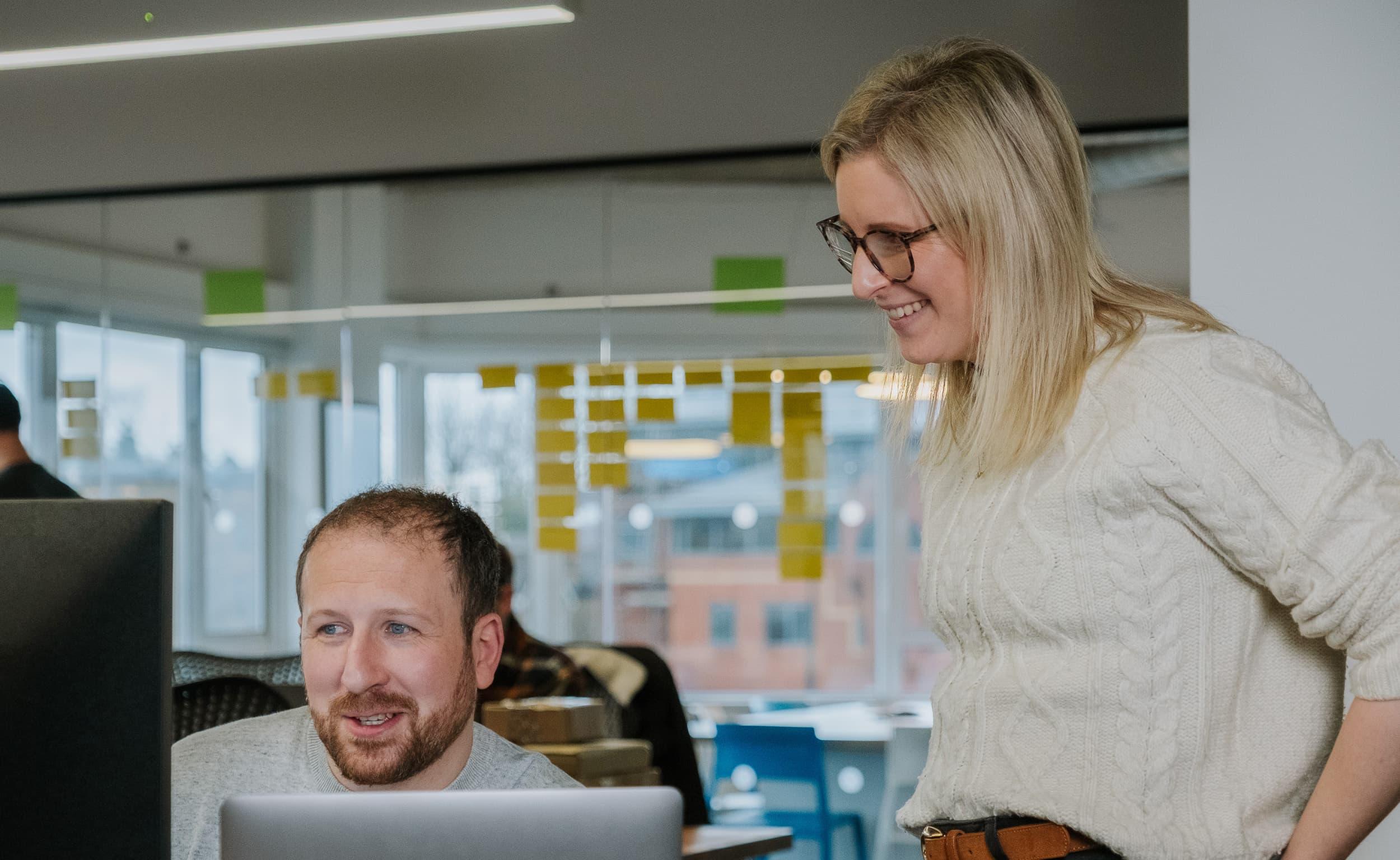 A woman stands beside a seated colleague, smiling as they review content together on a desktop computer in a bright office.