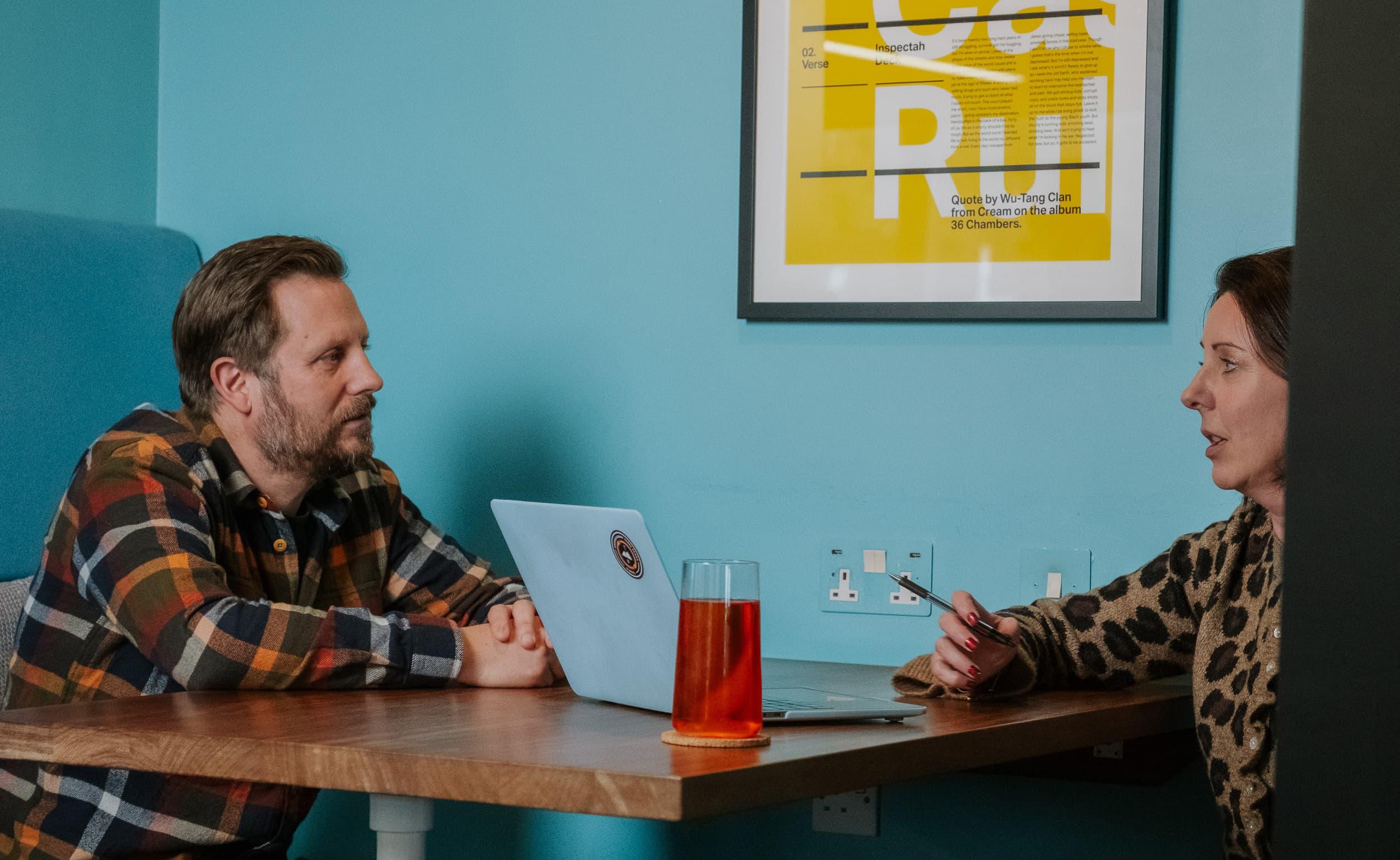 Two people sit across a table in a meeting room, talking through ideas with a laptop open between them and a glass of tea on the table