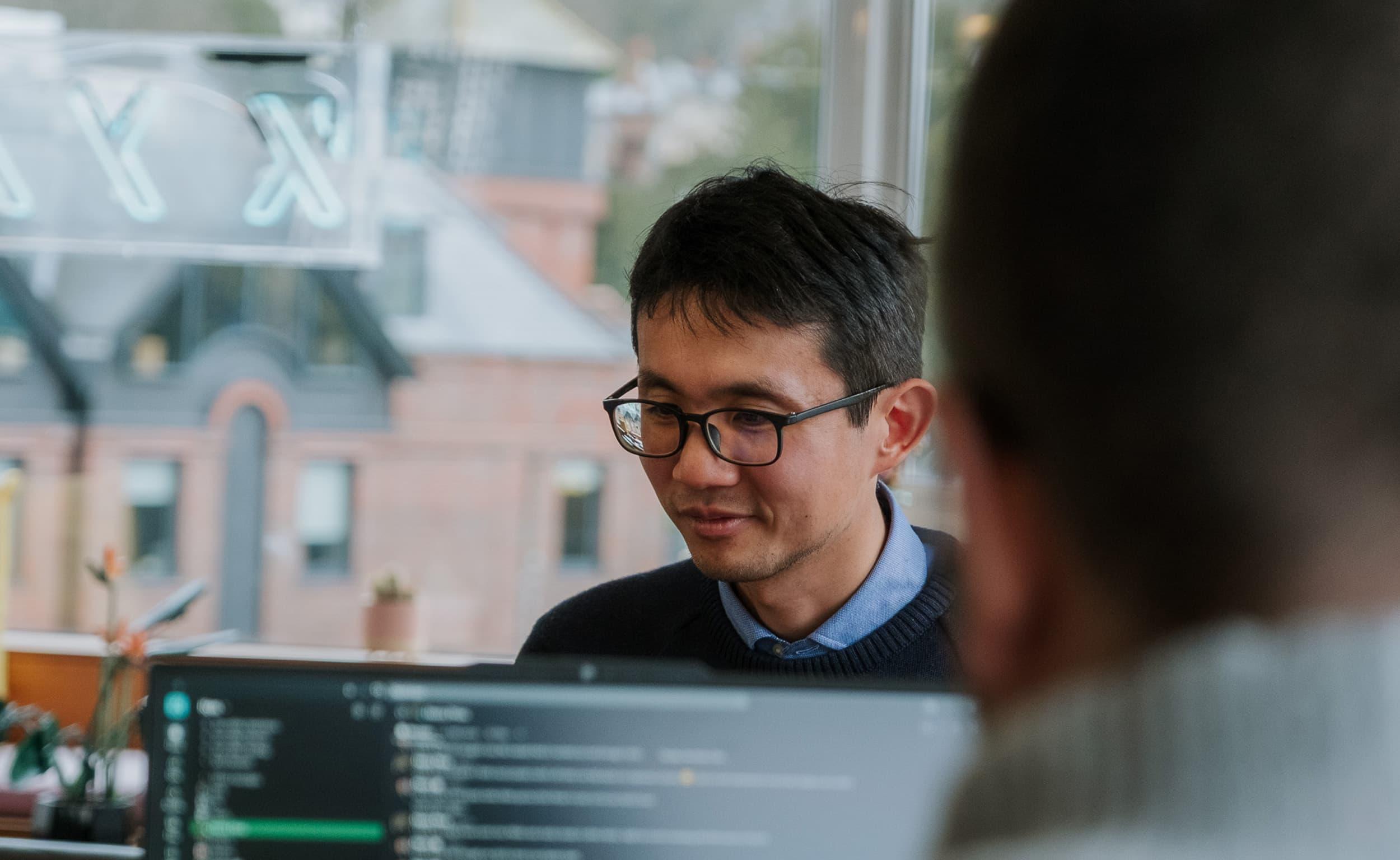 A man wearing glasses works at a computer in an office, with code visible on a screen in the foreground and colleagues blurred nearby.