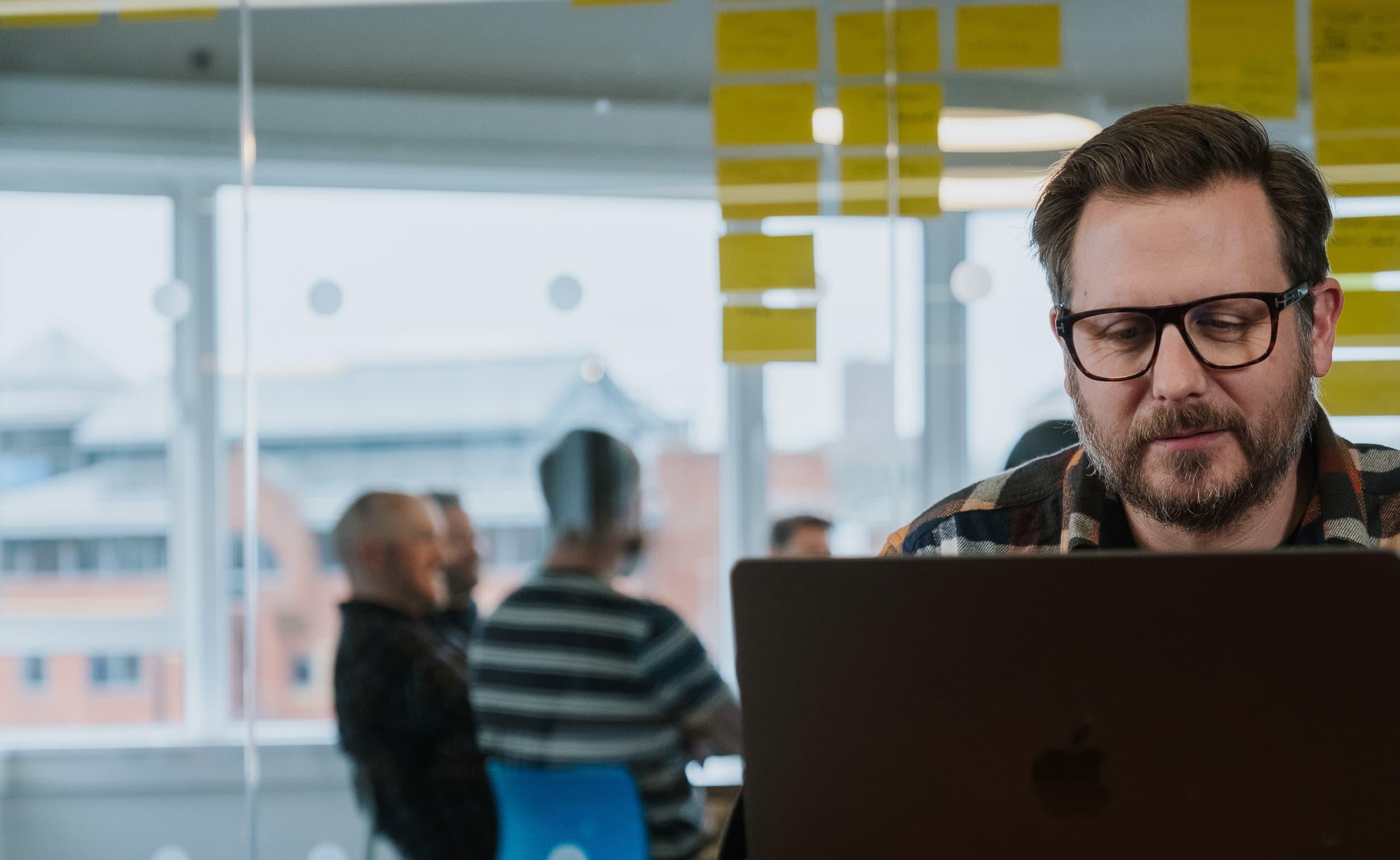 A man faces the camera working on a laptop in an office setting. In the background colleagues can be seen gathered in a meeting.