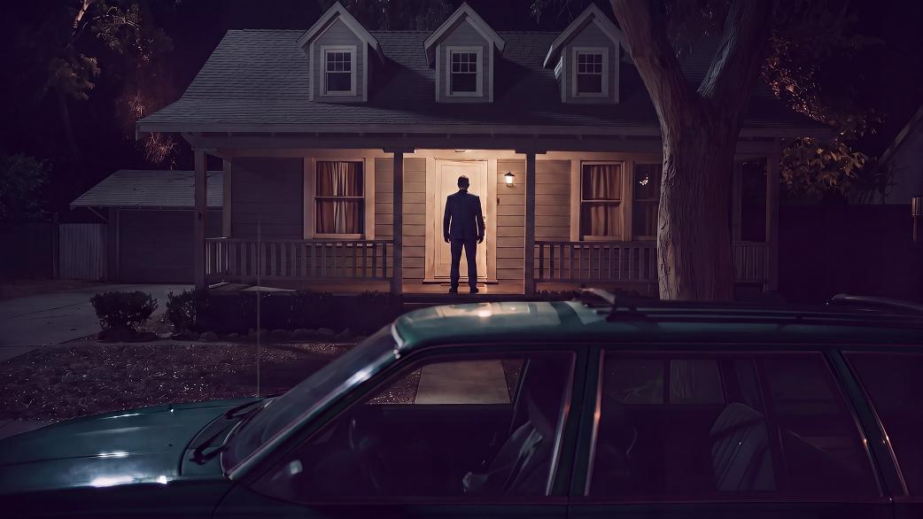 A cinematic night-time exterior: a figure silhouetted in the lit doorway of a suburban house, with a car in the foreground.