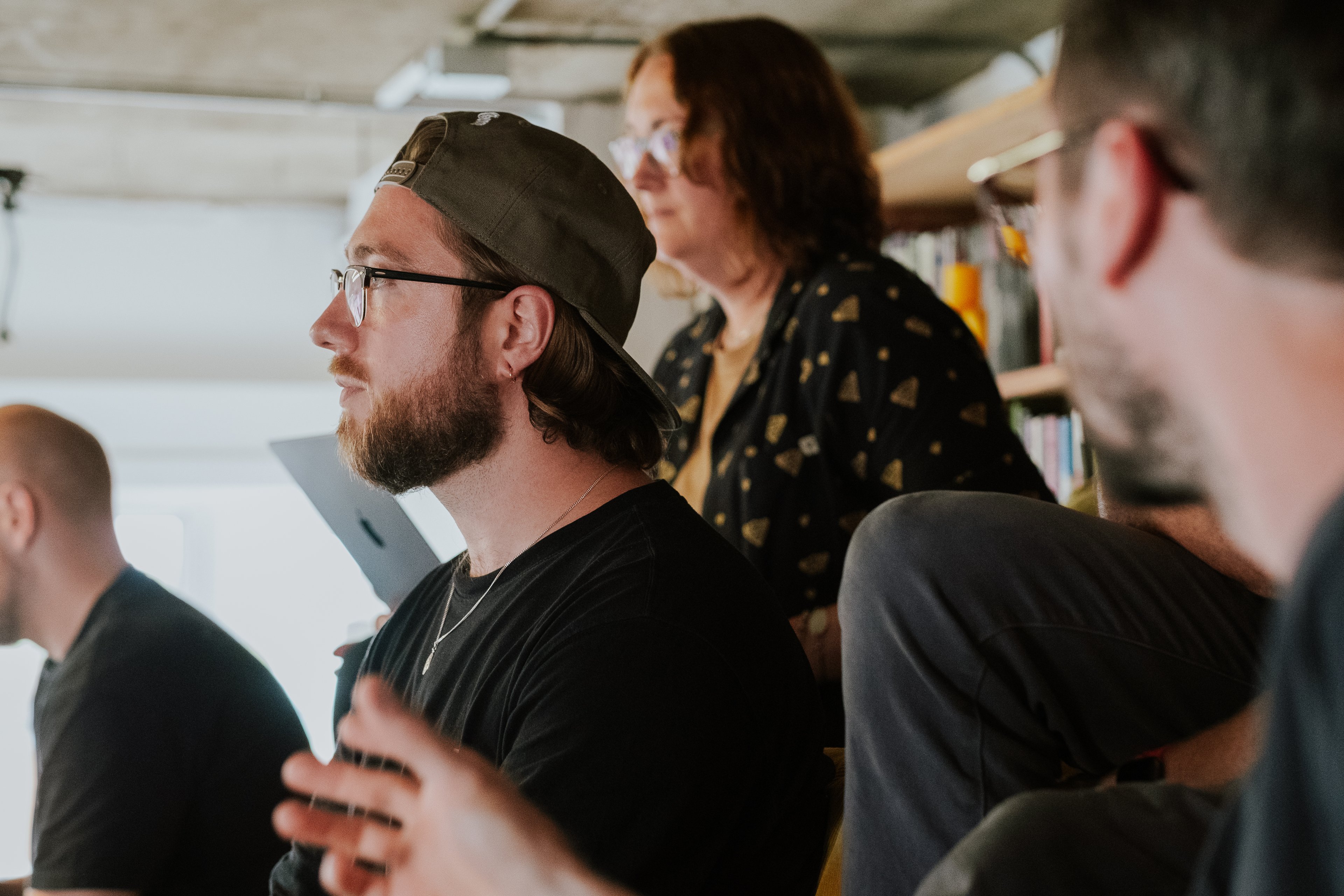 A man in a backwards cap and glasses listens attentively during a meeting, with two colleagues blurred in the foreground and background.