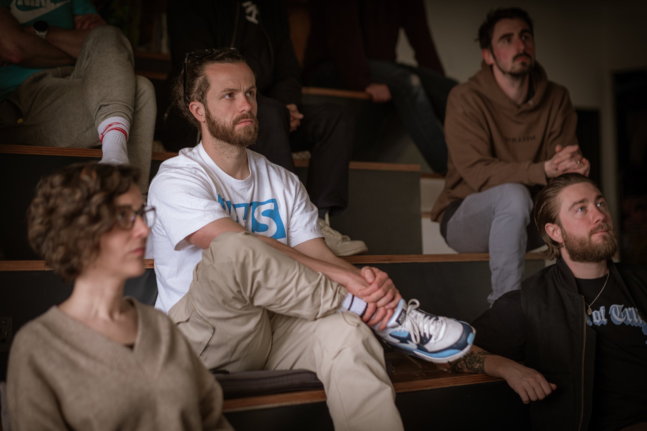 A group of people sit on indoor steps watching a presentation, with a man in an NHS T-shirt in the centre listening intently.