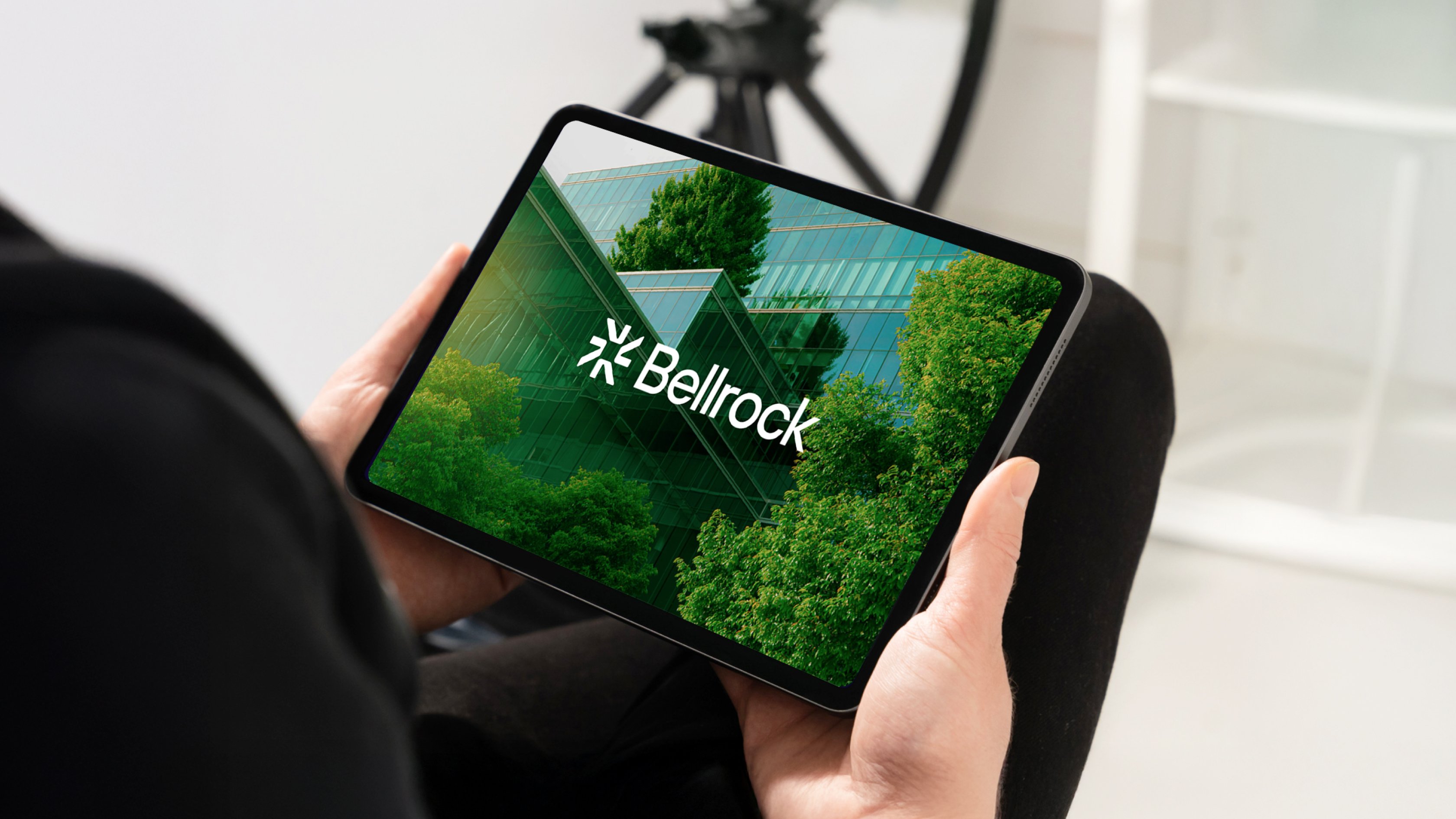 A person holding a tablet displaying the Bellrock logo over a photo of a modern glass building surrounded by green trees.