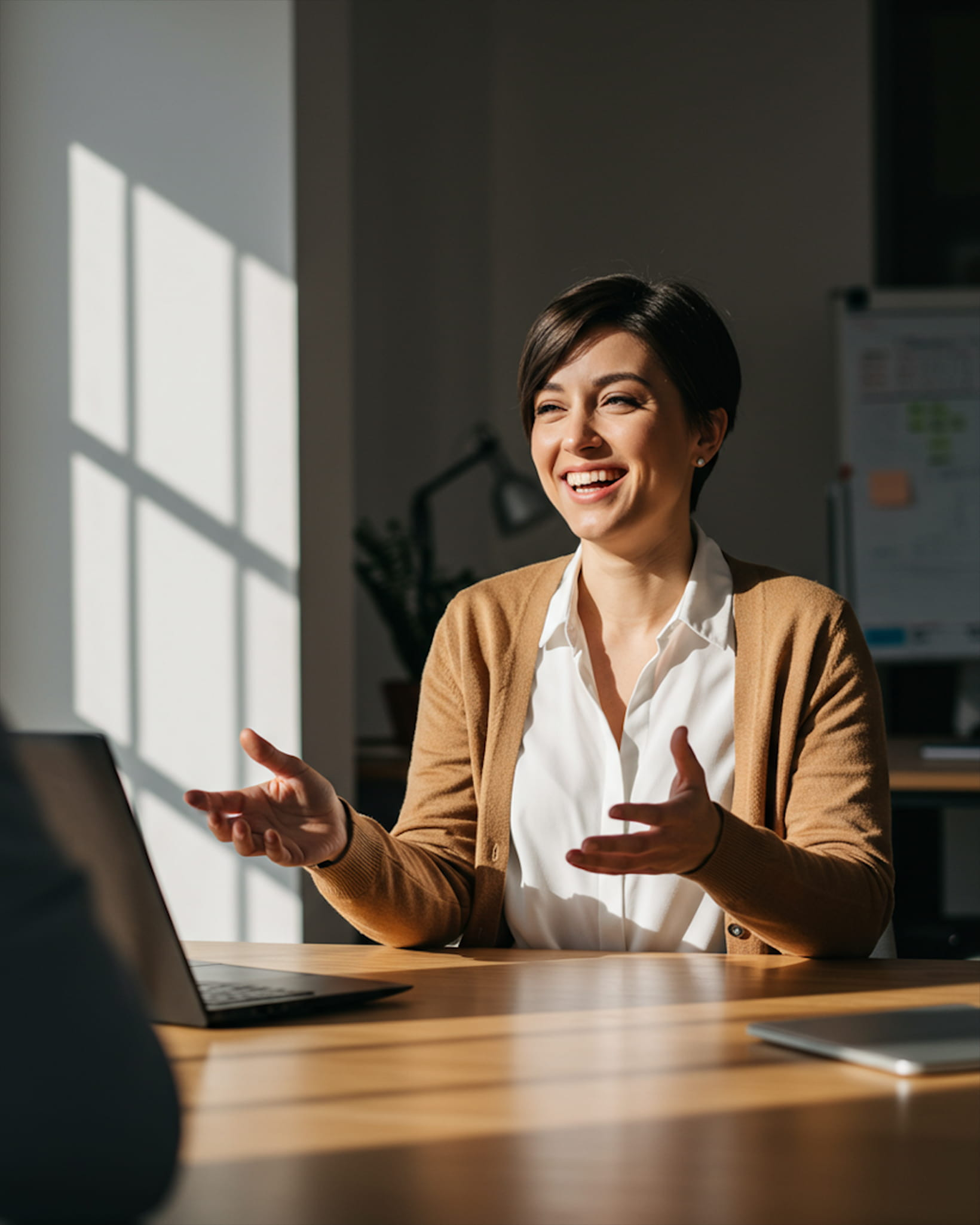 A woman smiling during a conversation at an office desk, gesturing with her hands while sunlight streams through a nearby window.