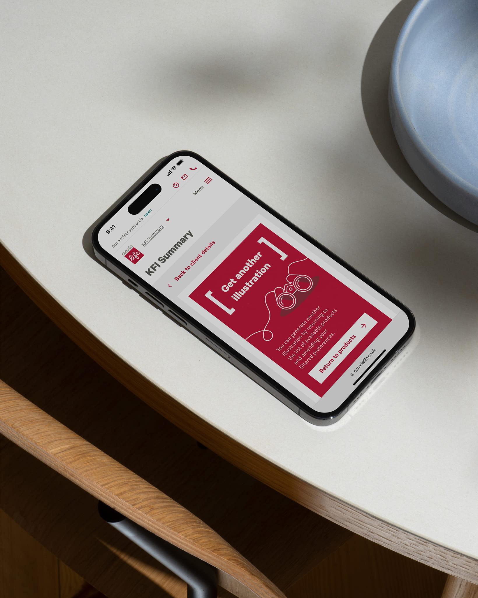 A smartphone resting on a table displaying a Canada Life KFI Summary page with a red card offering the option to get another illustration.