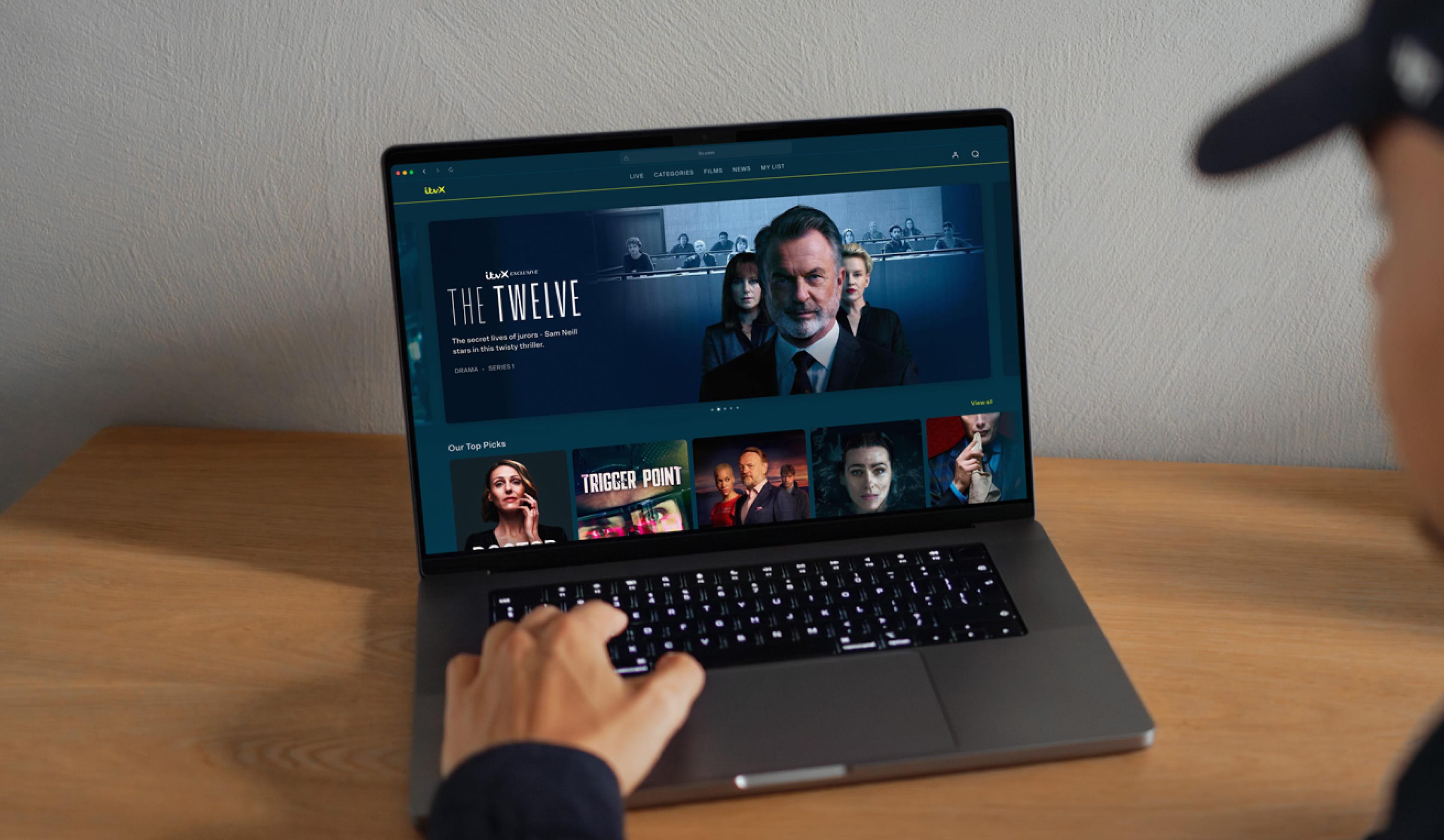 Person using a laptop at a desk, browsing the ITVX website with a featured banner for a drama series titled ‘The Twelve' and a row of recommended shows below.