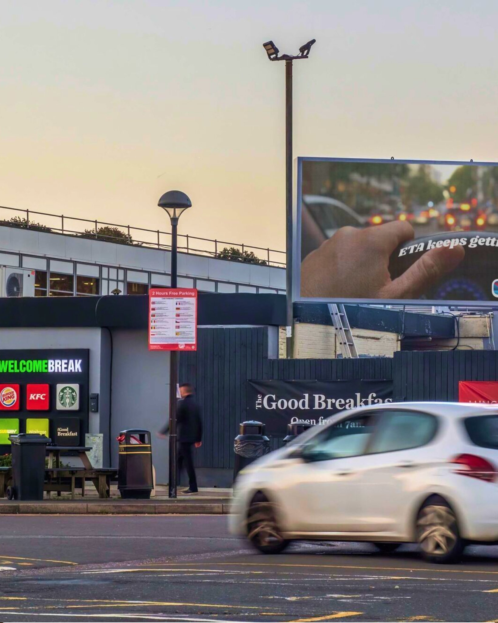 Roadside view of a Welcome Break service area with brand signs including Burger King, KFC and Starbucks, a large traffic billboard, and a white car driving past.