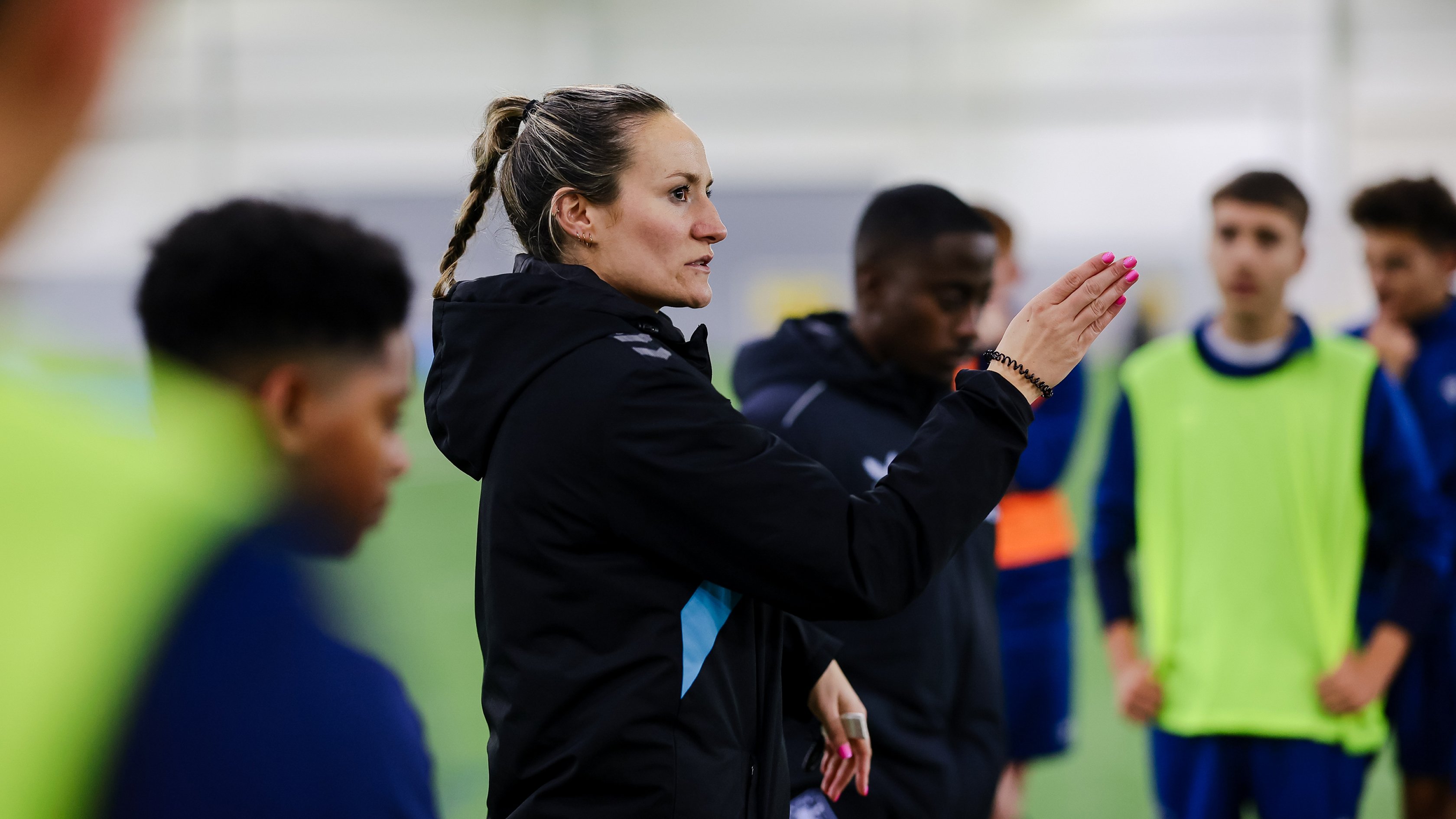 A football coach in a black jacket gestures while speaking to a group of young players during an indoor training session.