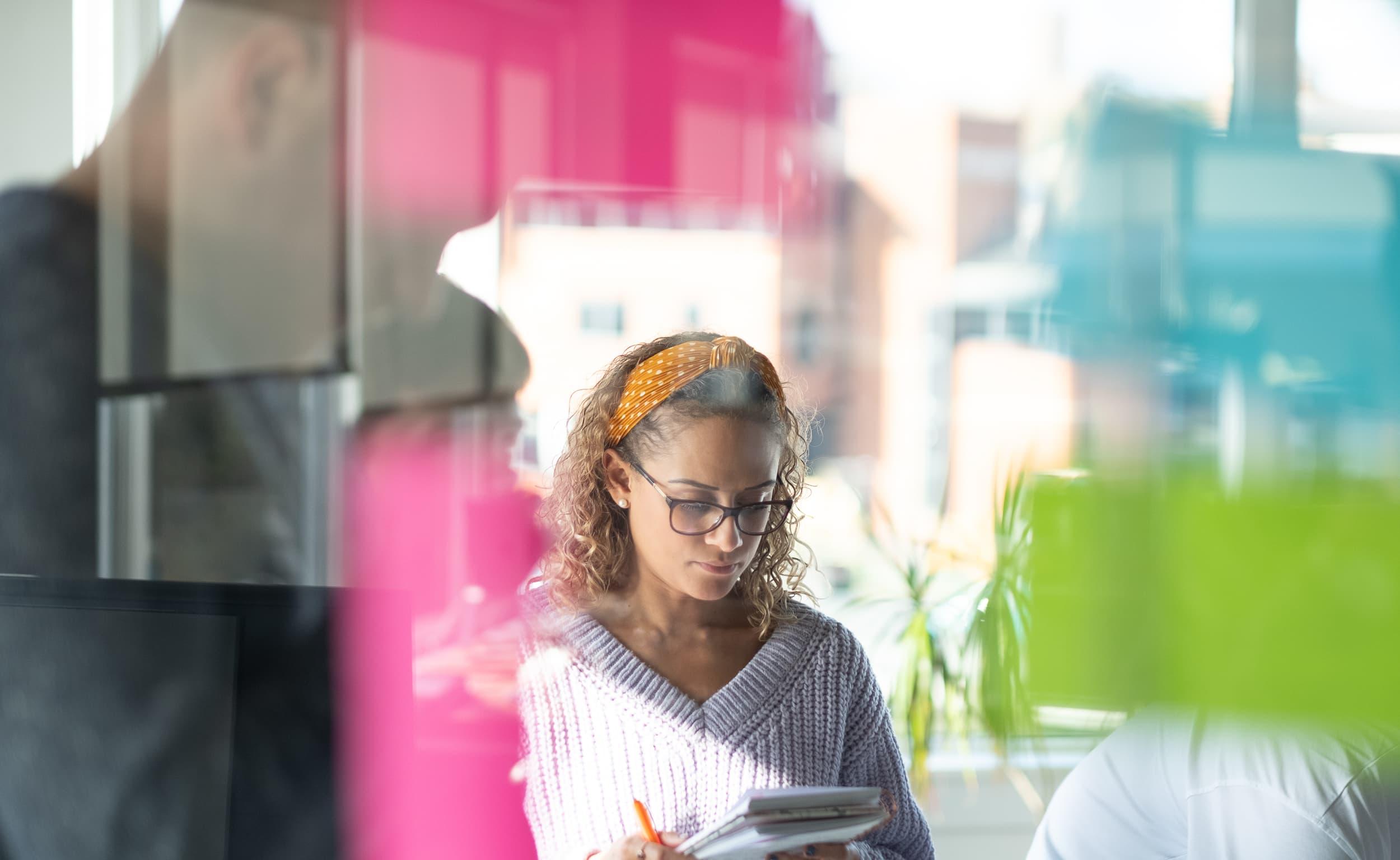 A woman with curly hair, wearing glasses and an orange headband, stands in a brightly lit office holding a notebook. Colorful sticky notes on glass panels are blurred in the foreground.