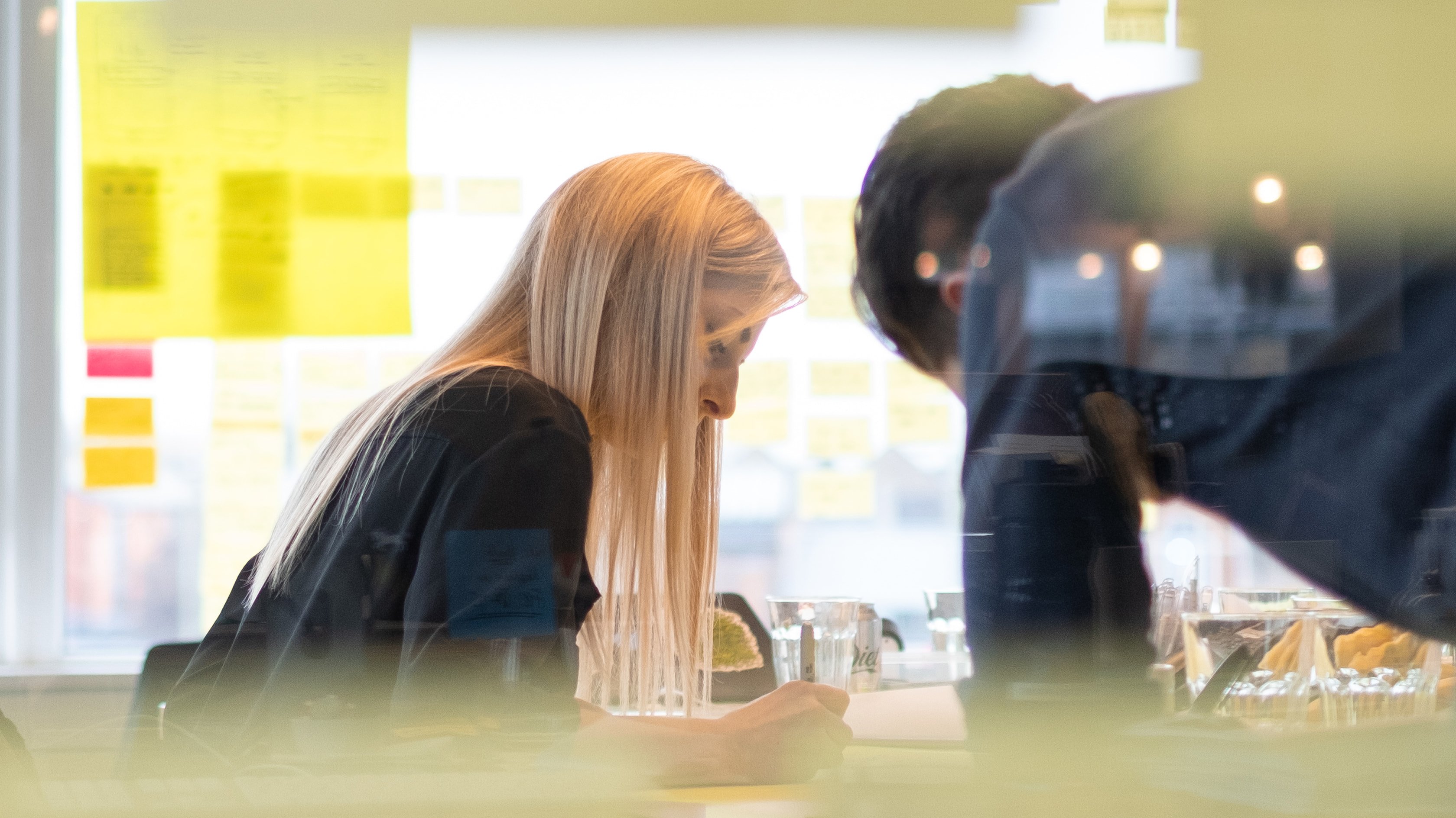 A close-up view of two colleagues writing notes during a workshop in an office setting. They are viewed through the glass wall of a meeting room, with soft reflections of light visible.