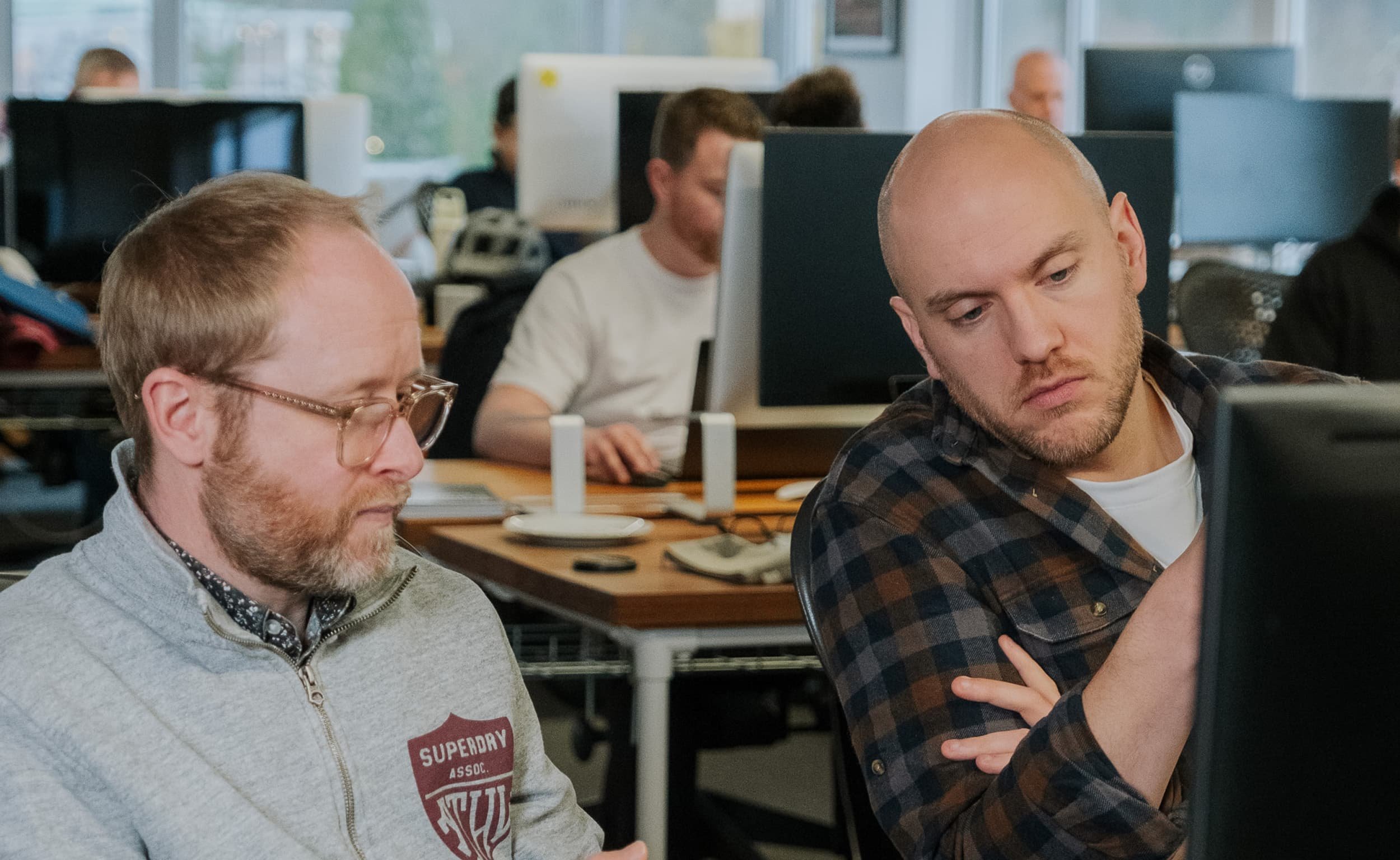 Two colleagues sit side by side at a desk in an open-plan office, focused on a computer screen while discussing work.