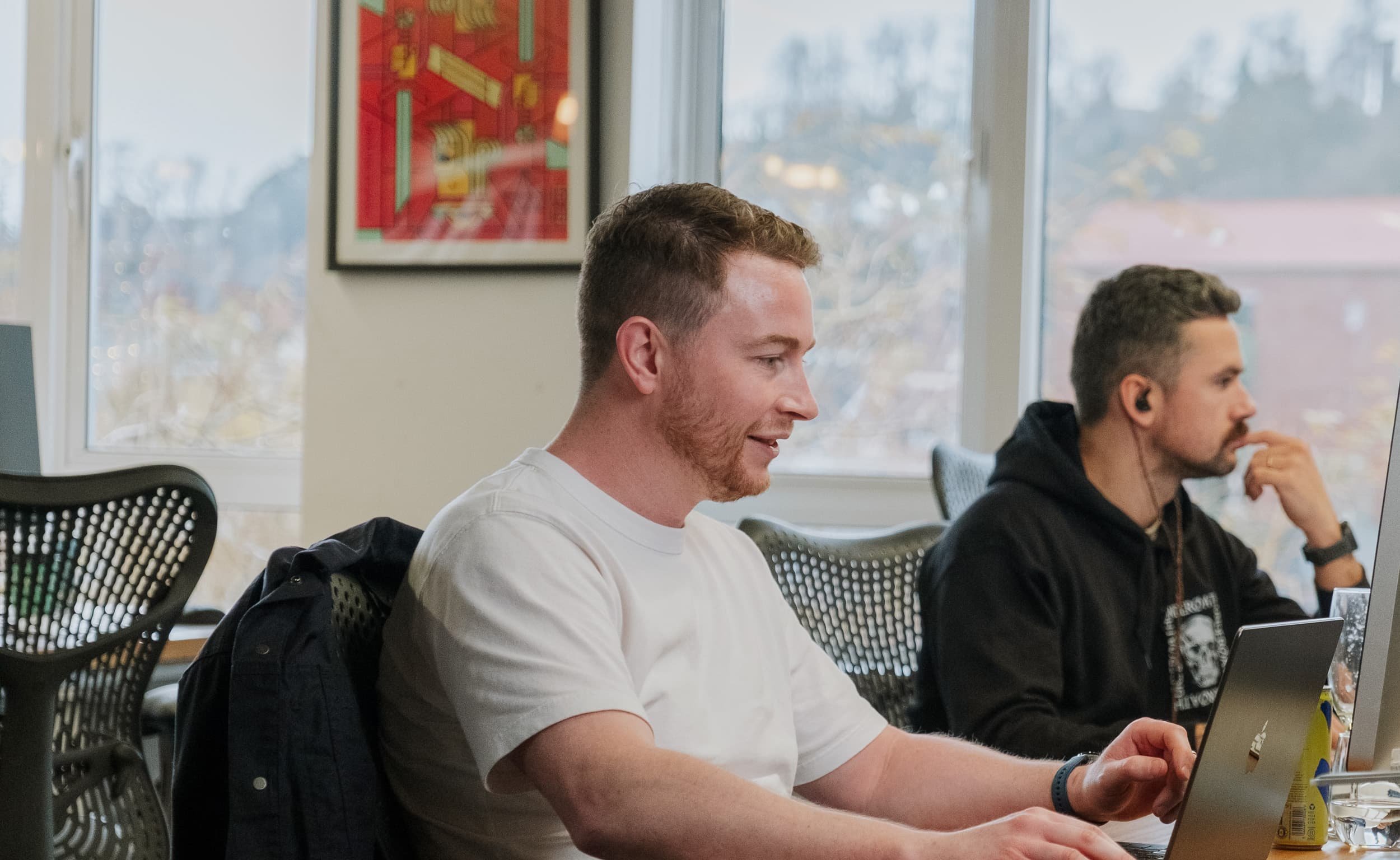 Two colleagues work at laptops in an open office, one in the foreground concentrating while another listens with earbuds in.
