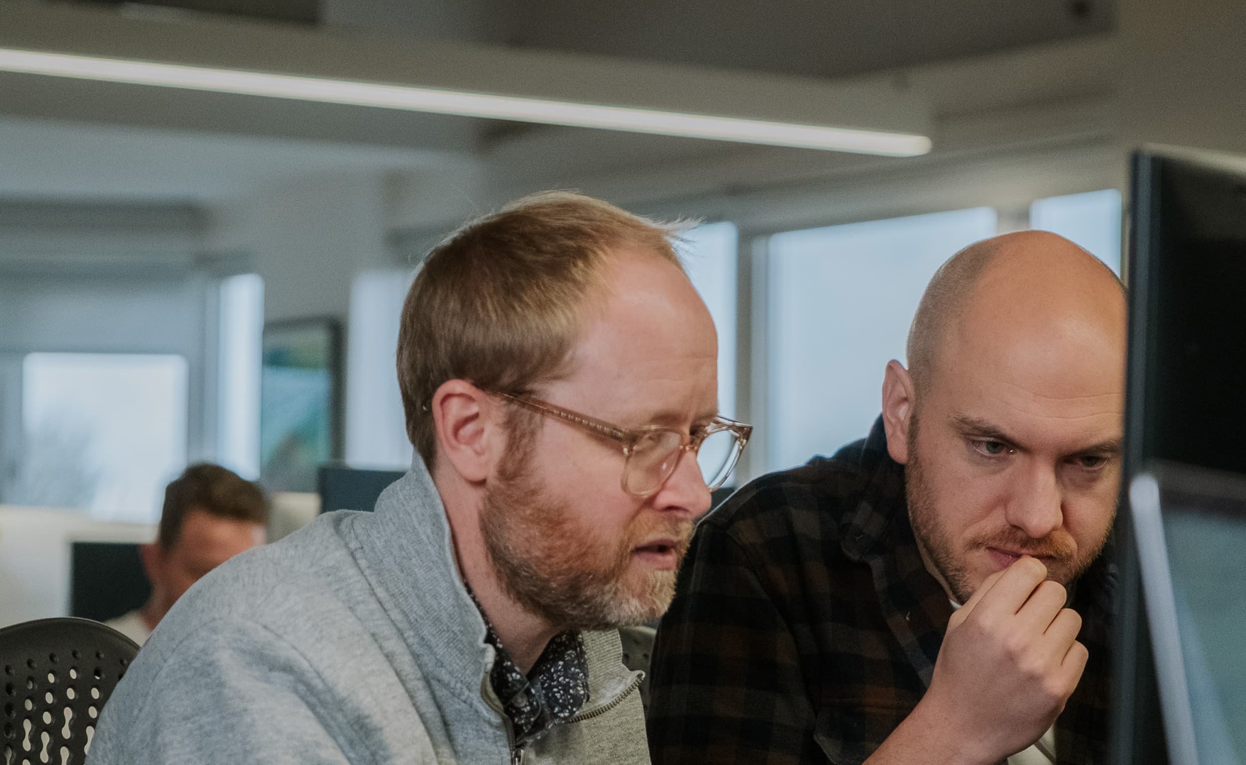Two men lean in toward a computer screen, concentrating as they review something together at a desk.