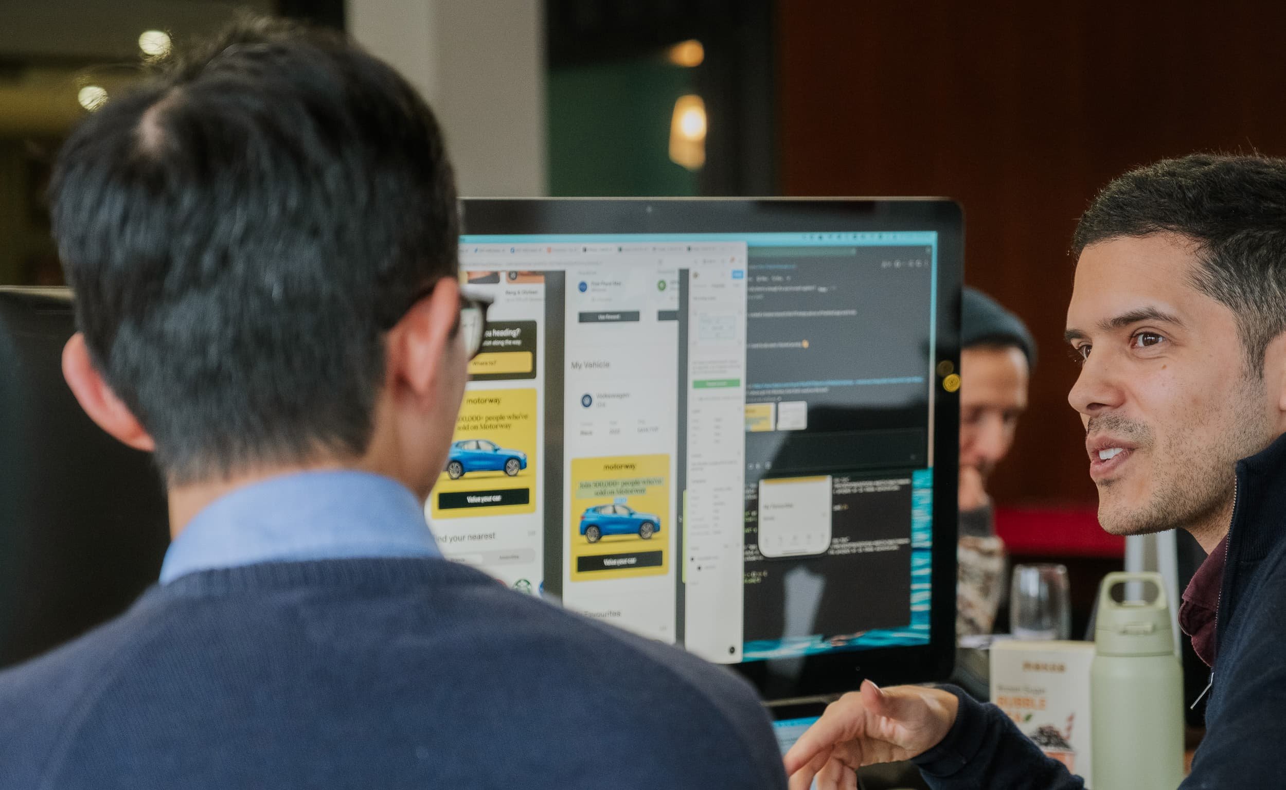 Two colleagues discuss a design on a large monitor, with interface screens and code visible as one gestures while explaining.
