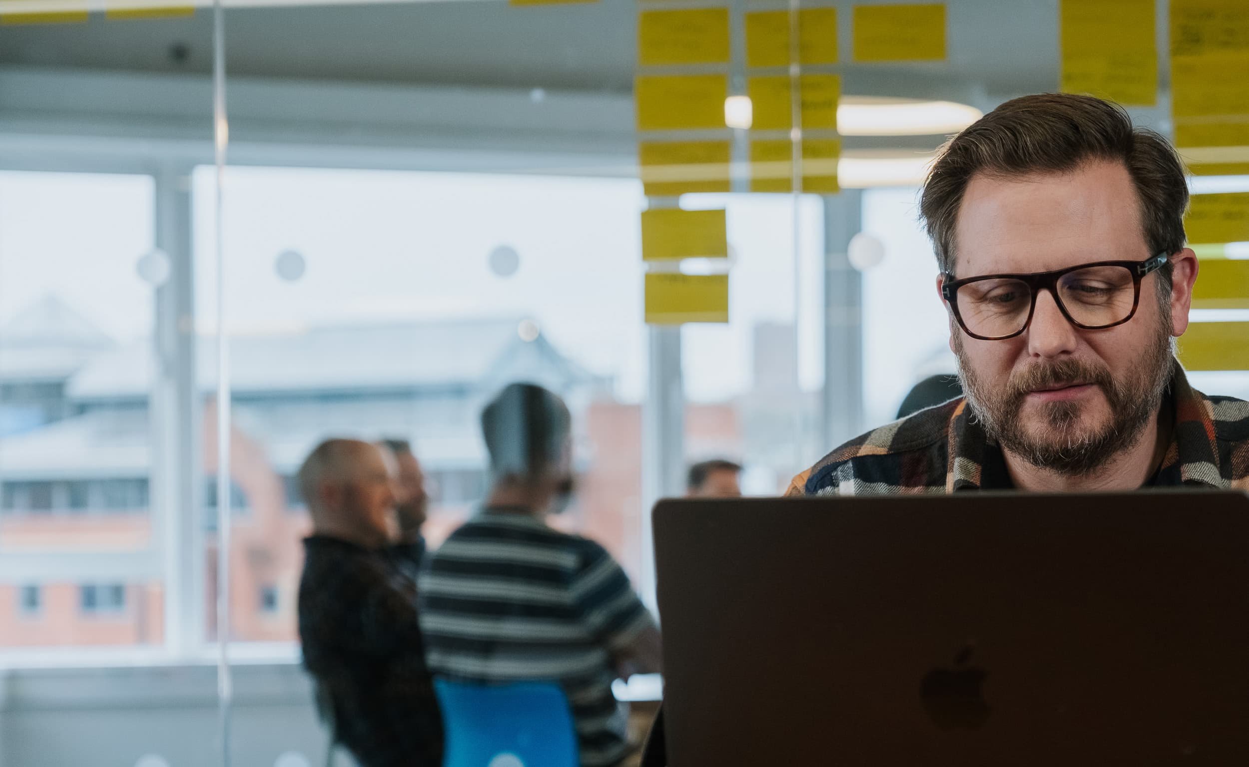 A man works on a laptop at a desk, with a glass wall behind him covered in yellow sticky notes and colleagues chatting beyond.