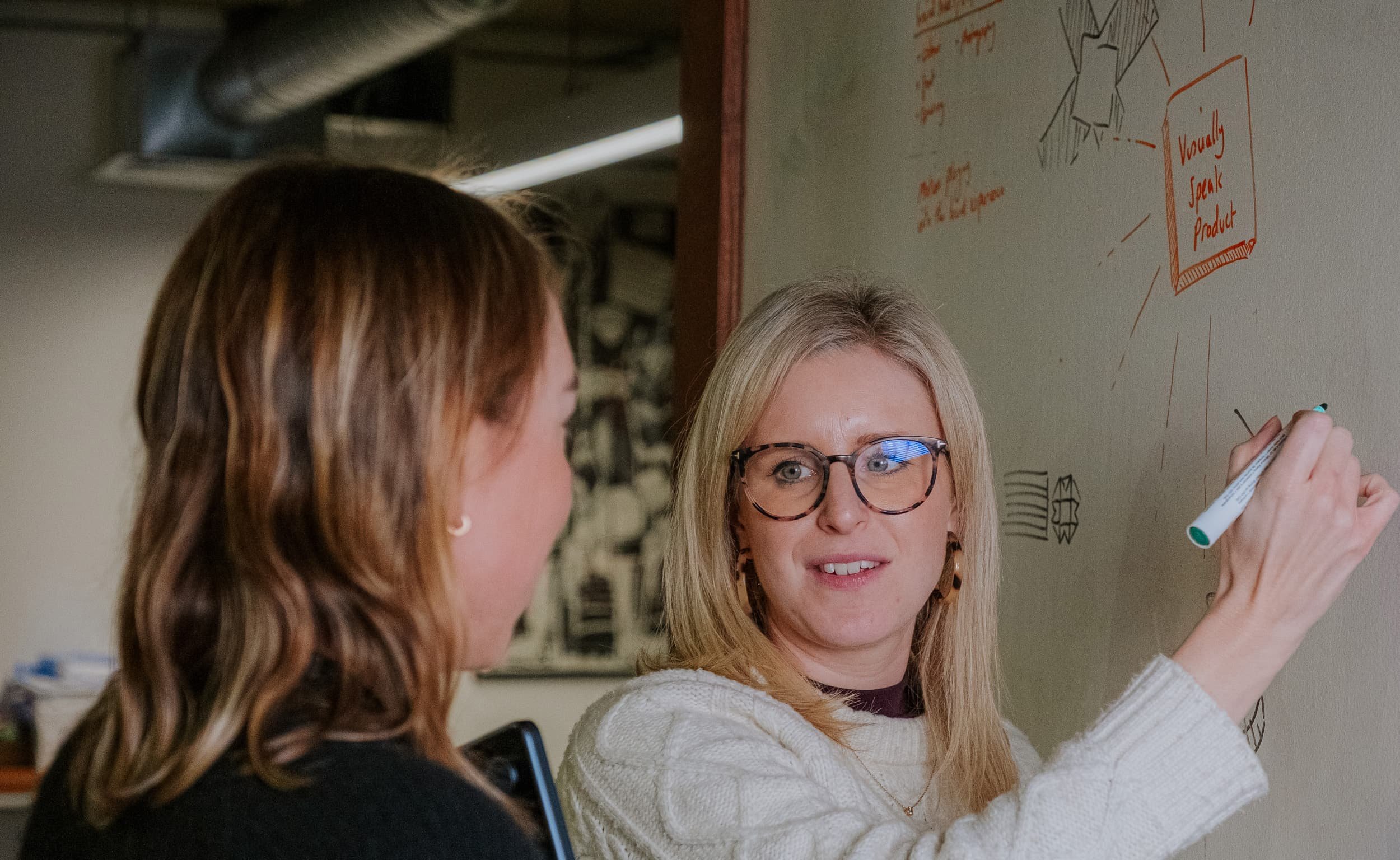 A woman writes on a whiteboard during a discussion, turning to speak with a colleague standing beside her.