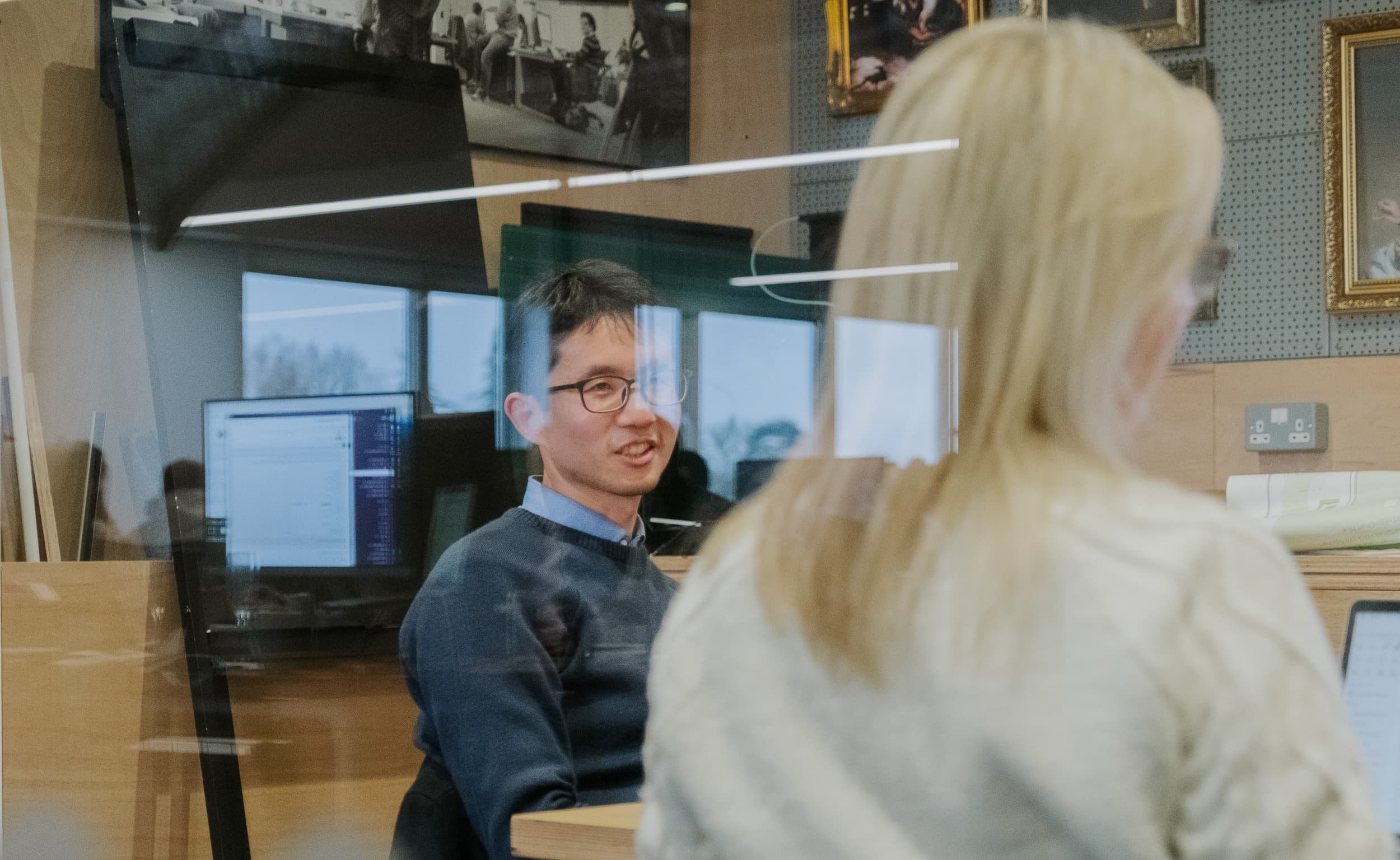 A man speaks during a meeting, seen through a glass wall with reflections of screens and the office space around him.