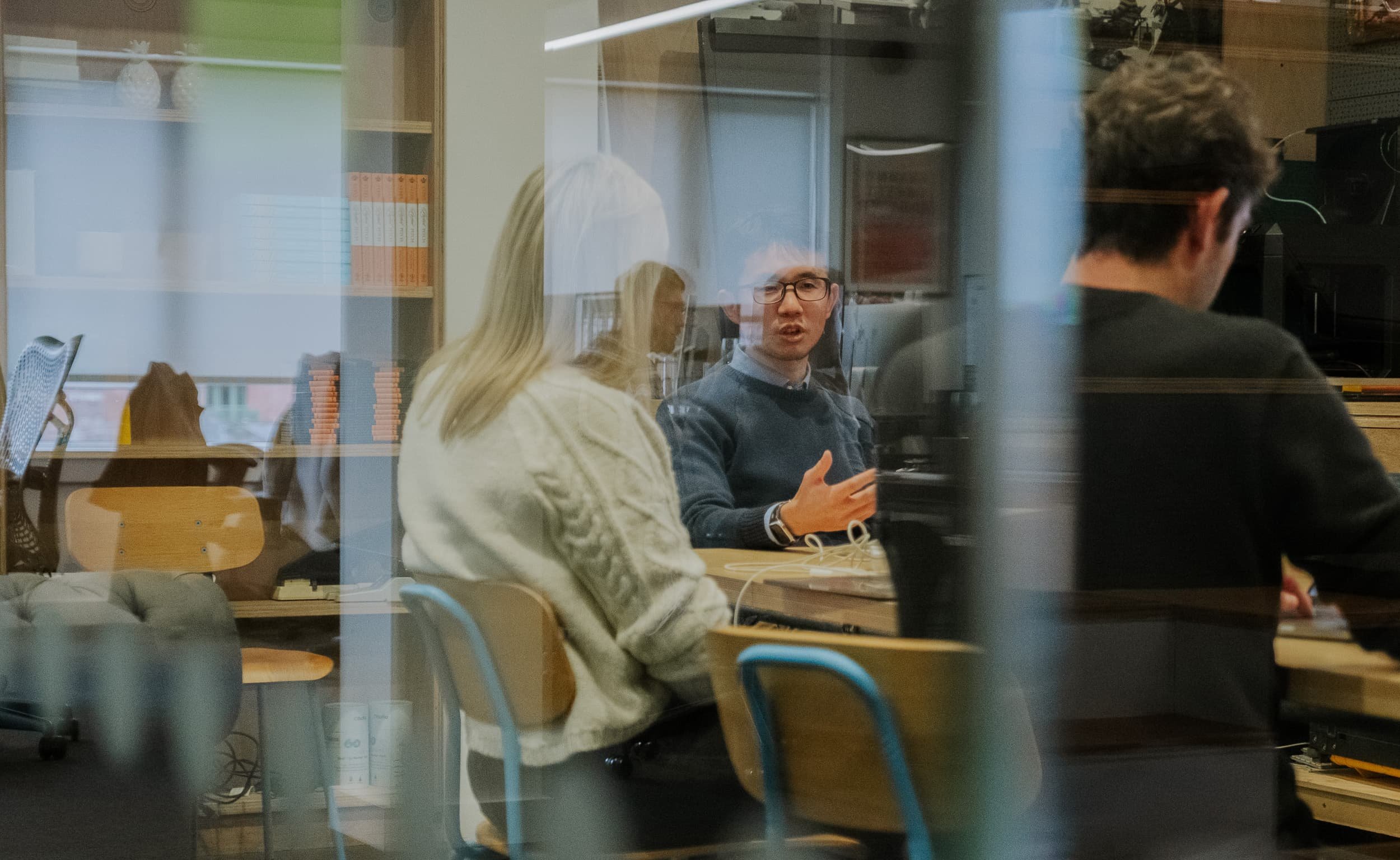Three colleagues sit around a table in a meeting room, seen through a glass wall, as one person gestures while explaining an idea.