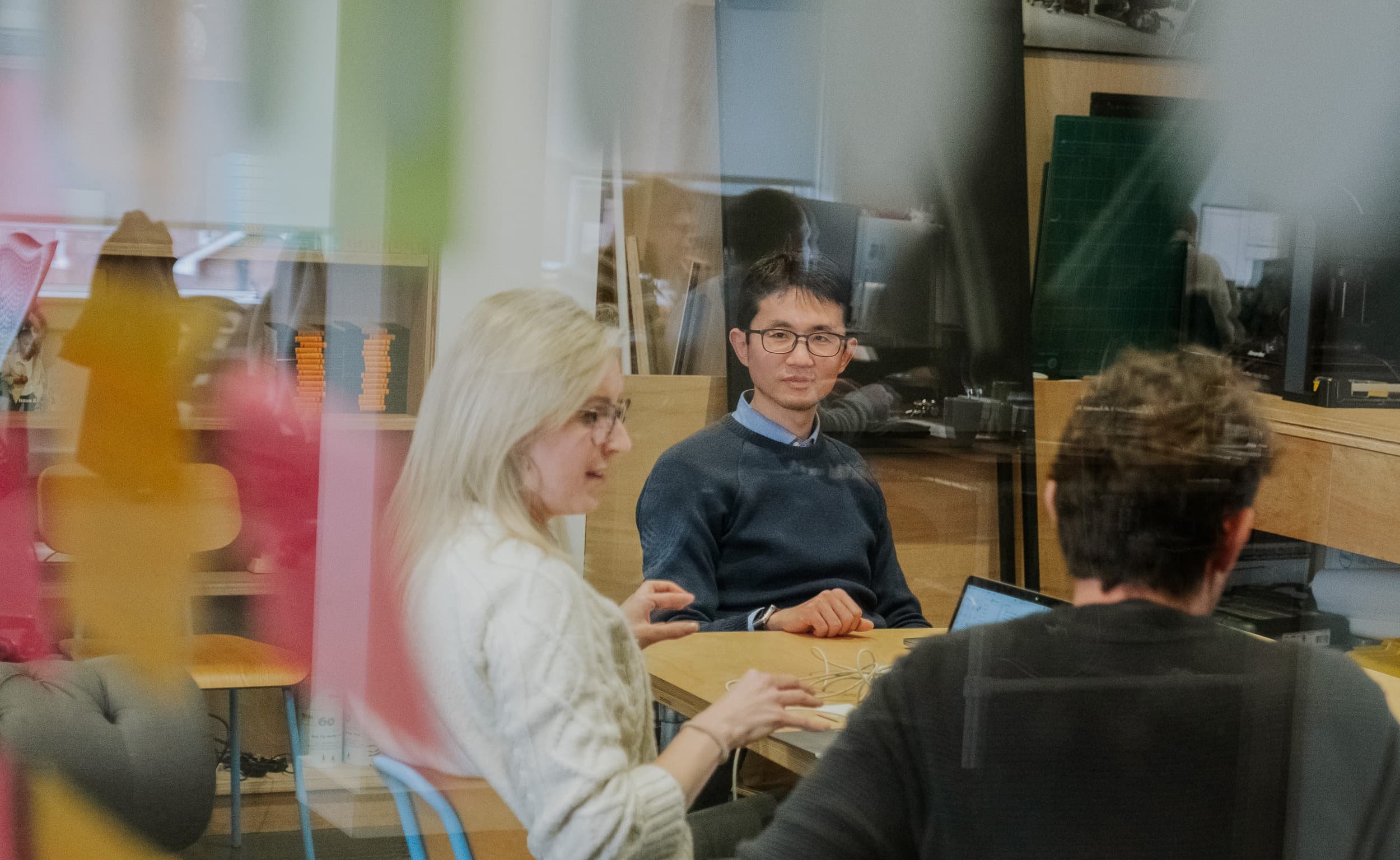 Colleagues sit around a table in discussion, viewed through reflections on a glass wall, with laptops and notebooks on the desk.