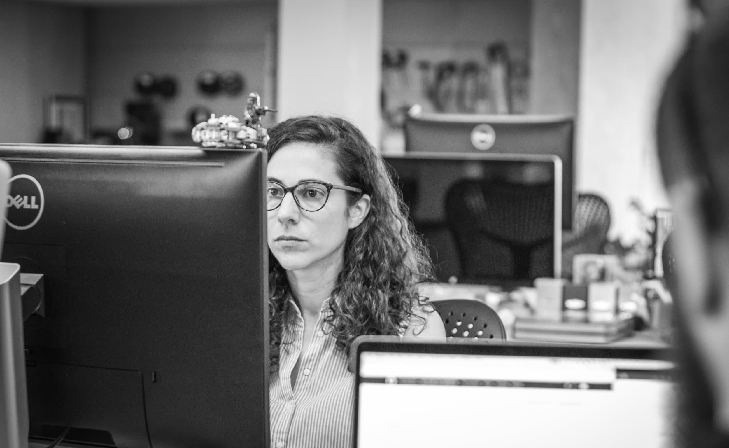 A woman wearing glasses works at a computer in an open plan office with other screens and desks in the background