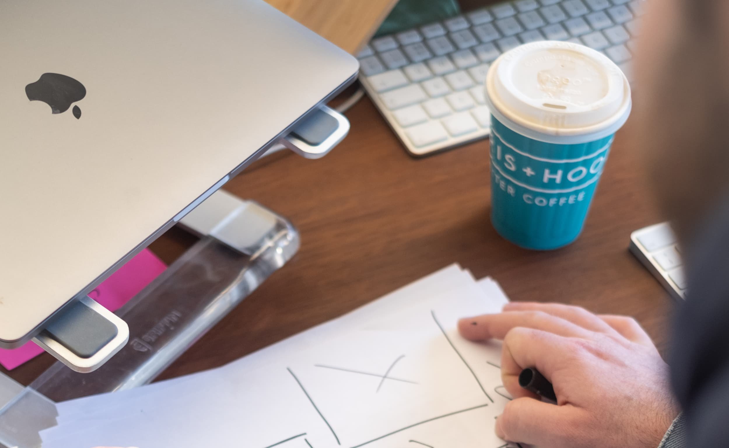 Laptop on stand, coffee cup, and hand sketching wireframes