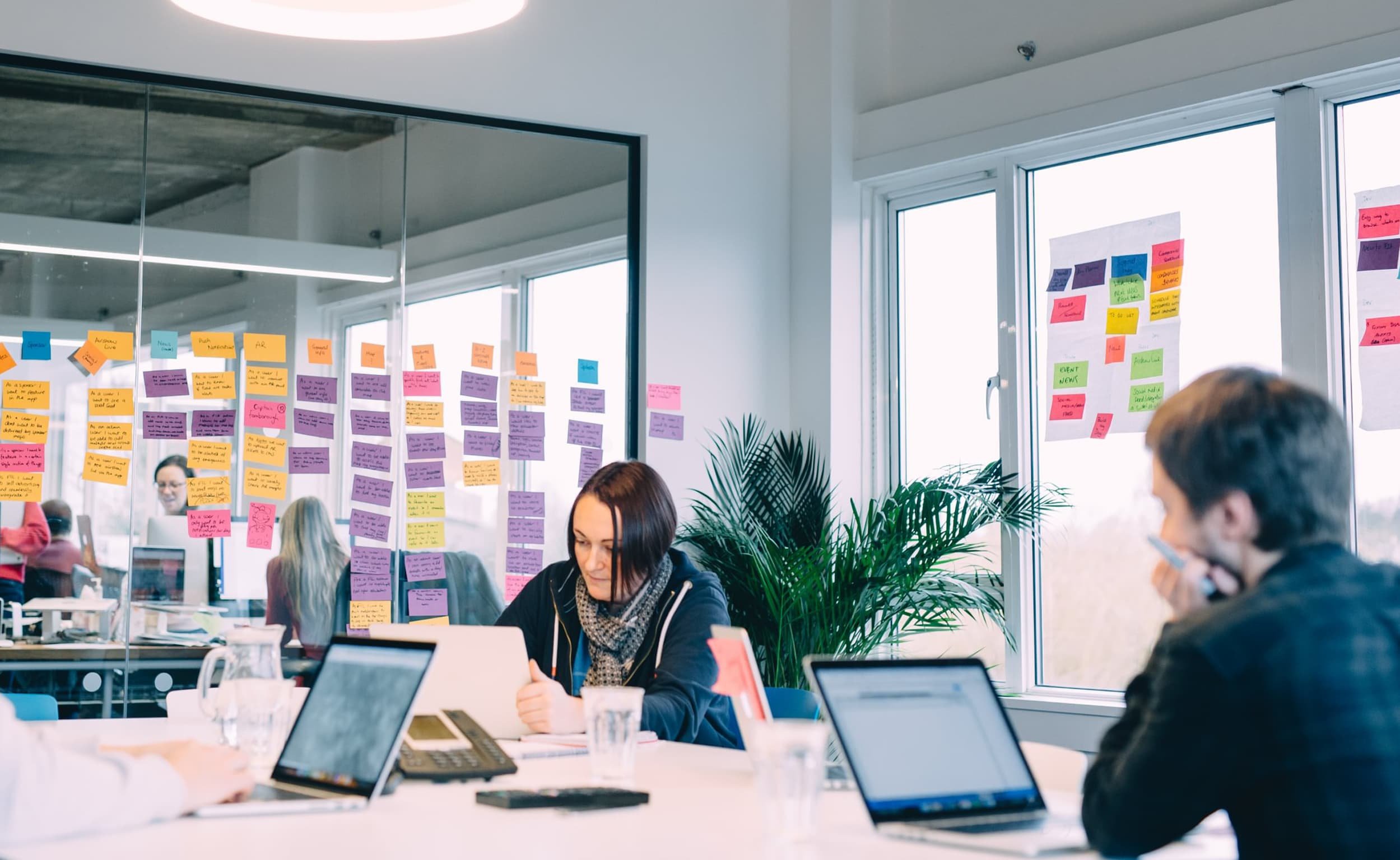 A group of people sit around a table in a meeting room, with large sections of the glass walls covered in colorful sticky notes. A woman with a laptop works at the table, while others brainstorm nearby.