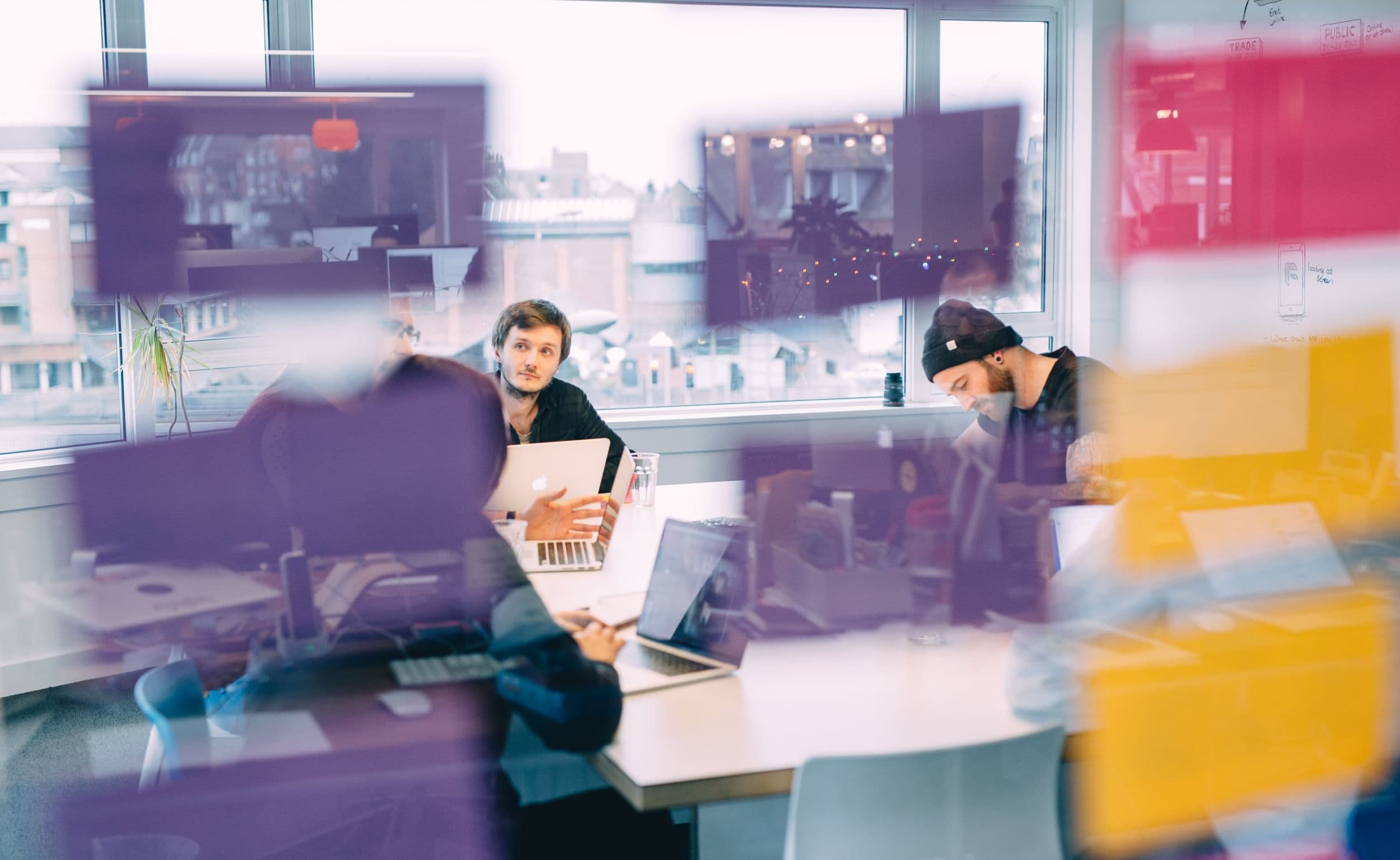 People sit around a meeting table in a modern office, collaborating with laptops. The scene is partially obscured by purple and yellow sticky notes on the glass wall.