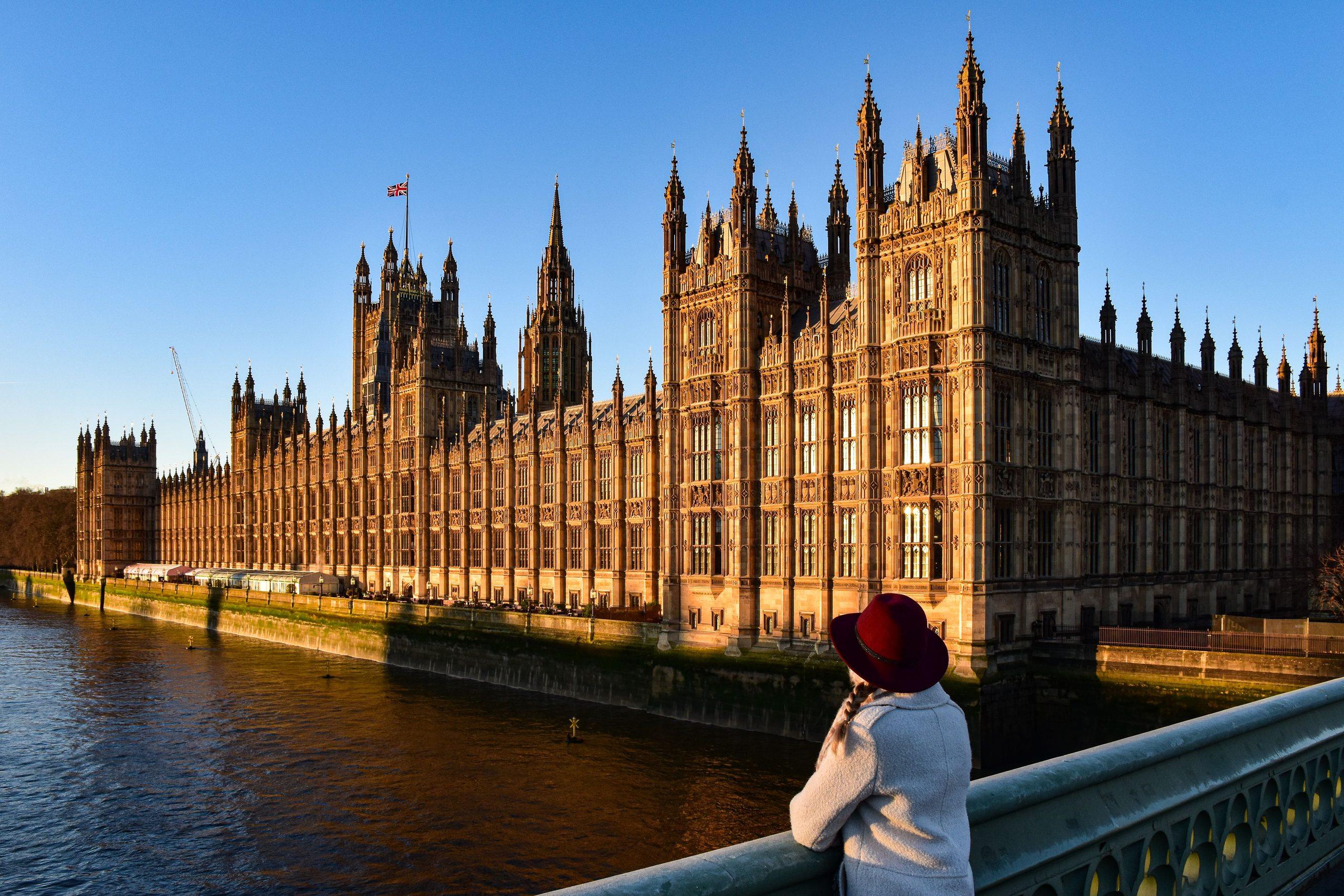 A person in a red hat looking across the River Thames towards the Houses of Parliament at golden hour.